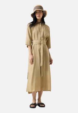 Kvinde iført en beige midi-kjole med bindebånd, brede ærmer, sorte slip-on sandaler, stribet stråhat og gyldne creoler, stående på hvid baggrund.