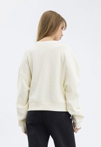 Felpa color crema con spalle scese e polsini a costine, caratterizzata da un orlo cropped e tessuto morbido e texturizzato; abbinata a jeans scuri.