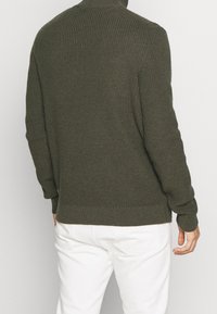 Olivgrüner, gestrickter Pullover mit hohem Kragen, strukturiertem Stoff, langen Ärmeln und geripptem Saum, kombiniert mit weißen Hosen.