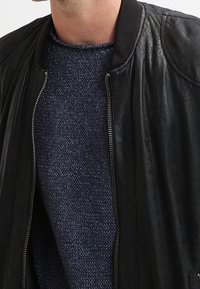 Svart läderbomberjacka med en ribbad krage, dragkedjestängning och slät textur. Bärs över en mörkblå stickad tröja.