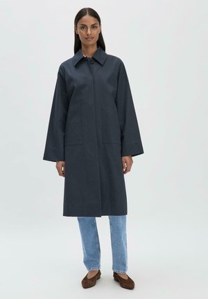 Cappotto blu scuro, alla lunghezza del ginocchio, con taglio dritto, colletto a punta, chiusura frontale con bottoni e due tasche applicate. Indossato sopra jeans azzurri.