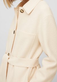 Abrigo beige con cinturón, confeccionado en tela suave, con cuello, dos bolsillos delanteros y botones en tono dorado. Vista lateral que resalta la textura suave y el ajuste.