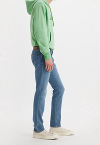 Personne portant un sweat à capuche vert clair, un jean bleu et des baskets beiges, debout de profil sur un fond uni.