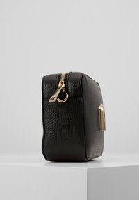 Piccola borsa di pelle nera testurizzata con cerniera e chiusura dorate, poggiata su un piedistallo bianco su uno sfondo grigio chiaro.