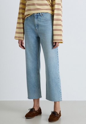 Persona che indossa jeans larghi corti azzurro chiaro, una camicia gialla a maniche lunghe a righe e mocassini marroni con lacci, in piedi su un pavimento semplice.