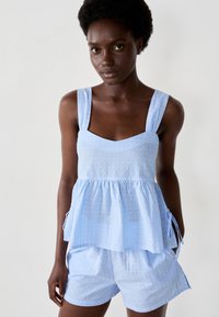 Top senza maniche di un blu chiaro con scollatura quadrata, tessuto texture e vita arricciata, abbinato a pantaloni corti leggeri dello stesso blu chiaro.