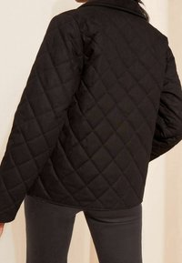 Černá prošívaná bunda s diamantovým vzorem, která má límec a volný střih. Textura je hladká, s dlouhými širokými rukávy.