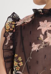 Sort gennemsigtig bluse med blomsterbroderi i pink og gul. Har flæseærmer og lodret syning. Letvægts, tekstureret stof.