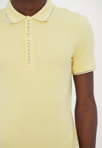 Světle žluté polo tričko vyrobené z měkké látky, s dvoupolohovým zapínáním a kontrastním šedým límečkem, s logem "Armani Exchange".