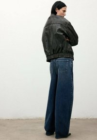 Chaqueta de cuero negra con un diseño corto y dobladillo elástico, combinada con pantalones de mezclilla oscuros de pierna ancha que cuentan con bolsillos traseros y un corte clásico.