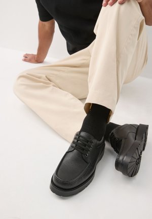Person trägt beige Hose und schwarze Socken, sitzt mit einem angewinkelten Bein und zeigt schwarze, strukturierte Leder-Schnürschuhe mit dicken Sohlen.