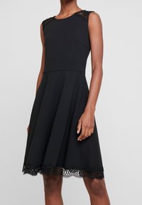 Czarna sukienka bez rękawów z dopasowanym gorsetem, rozkloszowaną spódnicą i koronkowymi akcentami na ramionach oraz u dołu, z fakturowanym koronkowym wykończeniem na końcu.