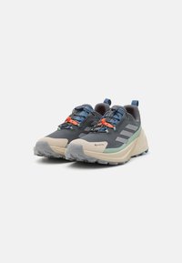 Buty biegowe z szarym wierzchem, zieloną i brązową podeszwą, wielokolorowymi sznurówkami, wyraźnym wzorem bieżnika i oznaczeniem GORE-TEX.