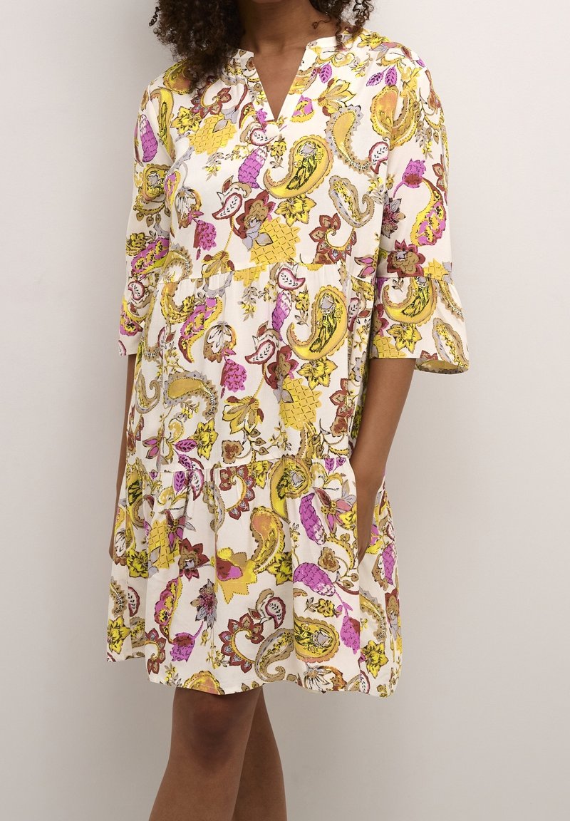 Femme portant une robe ample mi-longue blanche à motifs floraux paisley jaunes, roses et marron avec des manches trois-quarts.