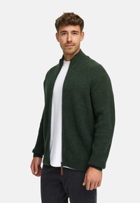 Dunkelgrüne, offen zu tragende Strickjacke mit hohem Kragen, gerippter Textur und braunem Lederreißverschluss, getragen über einem weißen T-Shirt und schwarzen Hosen.