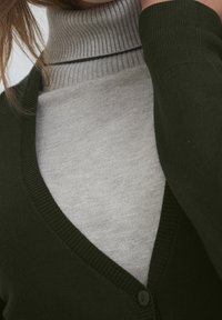 Tmavě zelený kardigan přes světle šedý žebrovaný rolák, s výstřihem do V a zapínáním na knoflíky, s jemnými texturami.