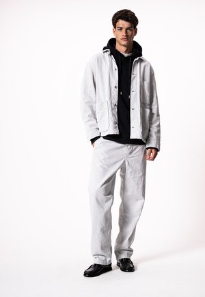 Jaunas vyras dėvi baltą švarką ir kelnes ant juodos kapišono, stovintis su rankomis kišenėse prieš baltą foną.