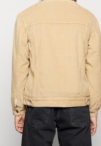 Manchesterjacka i ljusbeige, med vertikala ränder, justerbara manschetter med knappar och rak skärning. Bärs över mörka byxor.