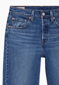 Jeans de mezclilla en azul medio, con un diseño clásico de cinco bolsillos, herrajes de cobre y detalles de costura visibles en la cintura y las costuras.