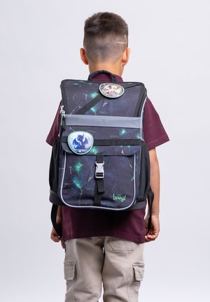 Junge trägt ein weinrotes Hemd und beige Cargohosen mit großem schwarzem und grünem Rucksack mit Drachen-Abzeichen, steht vor einfarbigem Hintergrund.