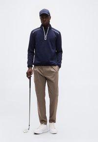 Άνδρας που φοράει μπλε σκουφί, μπλε πουλόβερ με φερμουάρ στο λαιμό και μπεζ παντελόνια, κρατώντας μια ράβδο γκολφ, στέκεται μπροστά σε λευκό φόντο.