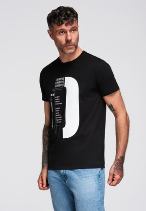 Mann mit Tattoos, der ein schwarzes Grafik-T-Shirt und blaue Jeans trägt und seitlich gegen einen schlichten hellen Hintergrund schaut.