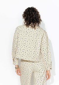 Veste beige cintrée avec un motif floral bleu marine, manches longues et poignets boutonnés. Assortie avec un pantalon à motif assorti.