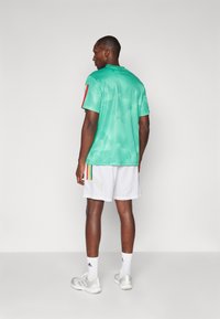 Camisa desportiva em malha verde com detalhes em riscas vermelhas e multicoloridas, combinada com calções brancos com um detalhe de risca diagonal.