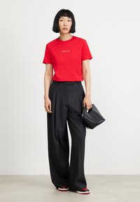 Red cotton t-shirt with white text, high-waisted black trousers, and a small black handbag with a rounded shape. Flat footwear with a color accent.
