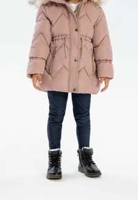 Veste rembourrée rose avec capuche et bordure en fausse fourrure, présentant des motifs en diamant, une taille à cordon de serrage et des poches zippées. Associée à un jean sombre et des bottes noires.