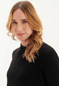 Pull en tricot noir avec une texture et un col rond. Cheveux ondulés et mi-longs, encadrant le visage sur un fond neutre.