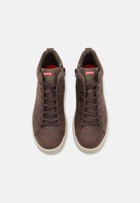 Brown suede high-top sneakers with perforated toe, cream rubber sole, and contrasting red and green branding on the tongue.