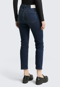 Naine, kes kannab tumedat sinist teksapükse ja musti loafer'eid, seisab seljaga kaamera poole, heleda halli tausta ees.