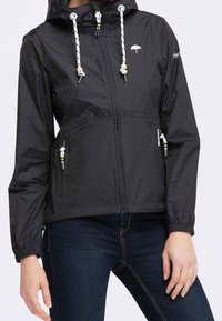 Giacca a vento nera con cappuccio, dotata di lacci bianchi e tasche con zip, indossata su jeans blu scuro.