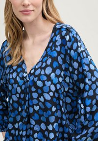 Blusa blu a fantasia di cuori con scollatura a V e maniche 3/4. Il tessuto ha una texture liscia e uno sfondo scuro.