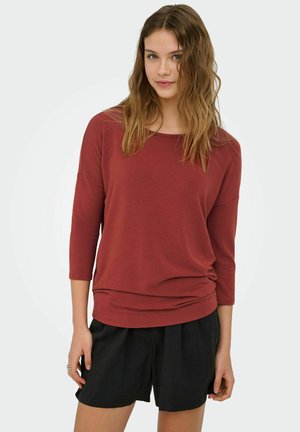 Młoda kobieta o falowanych brązowych włosach, ubrana w ceglastoczerwony długi rękaw i czarne szorty, stojąca na jednolitym białym tle.