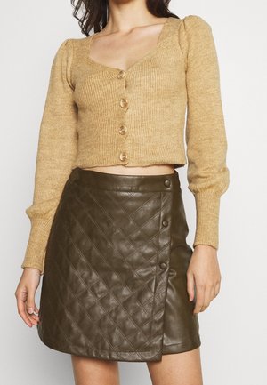 Frau trägt einen kurz geschnittenen, beigefarbenen Strickpullover mit Knöpfen und einen gesteppte dunkelolivgrüne Leder-Minirock mit Knopfdetails.