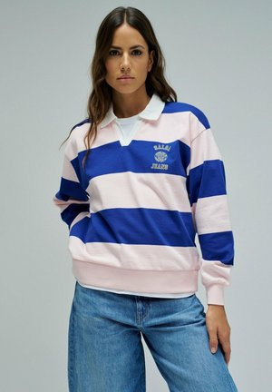 Mujer que lleva una camiseta de rugby de manga larga con rayas horizontales azules y rosas y cuello blanco, combinada con vaqueros azules.