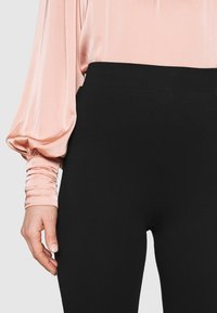 Svarta högmidjade leggings med en slät yta, tillsammans med en ljusrosa satinblus med långa ärmar som har rynkade ärmdetaljer.