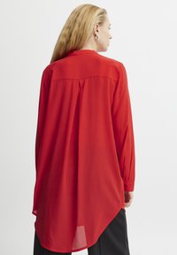 Blusa rossa a maniche lunghe con orlo curvo, caratterizzata da un leggero lucido e design con plissettatura sul retro. Il tessuto sembra leggero e morbido.