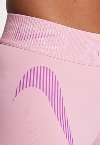 Rosa treningstights med ribbestrikket midje. Har et bølgete mønster i mørkeblått og lyselilla. Glatt tekstil med lett glans.