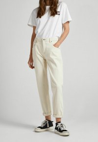 Kermanväriset korkeavyötäröiset farkut suoran lahkeensuukiristys, käänteet lahkeissa ja etutaskut, yhdistetty valkoiseen t-paitaan ja mustiin lenkkareihin.