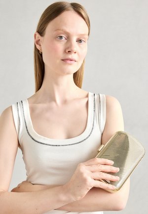 Ung kvinde med glat lysebrunt hår iført en hvid tanktop, der holder en metallic gylden clutch-pung mod en ensartet grå baggrund.