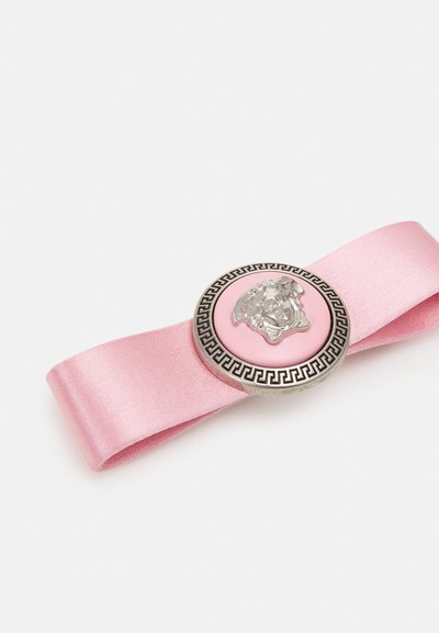 Nastro in satin rosa con un medaglione metallico rotondo che presenta una testa di leone argentata e un bordo geometrico nero.