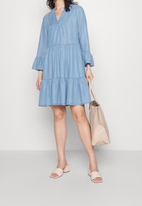 Žena stojí v světle modrých šatech ve vrstvách, drží béžovou tašku. Má na sobě světlé sandály na podpatku a má vlnité vlasy.