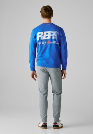Man in een blauwe sweatshirt met witte tekst en logo's, grijze joggerbroek en witte sneakers, staande tegen een effen grijze achtergrond.