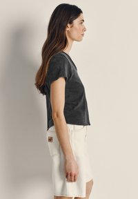 Naine mustas lühikeste varrukatega särk ja valge põlvikuni seelikuga seisab küljele pööratuna beeži seina ääres.