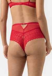 Roter Spitzen-String mit gewelltem Rand, transparentem Mesh-Einsatz am Rücken und einem Ausschnitt an der Taille, der filigrane Muster hervorhebt.