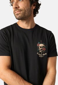 Zwarte katoenen t-shirt met kleurrijke borduursels van een schedel en roos op de linkerkant van de borst. Korte mouwen met een ronde hals.