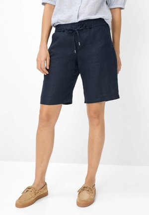 Person iført marineblå snørede shorts, lyseblå bluse og beige casual snøresko, stående mod en hvid baggrund.